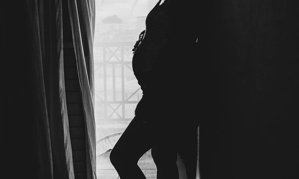 Silhouette of unrecognizable pregnant woman standing near window at home .pexels