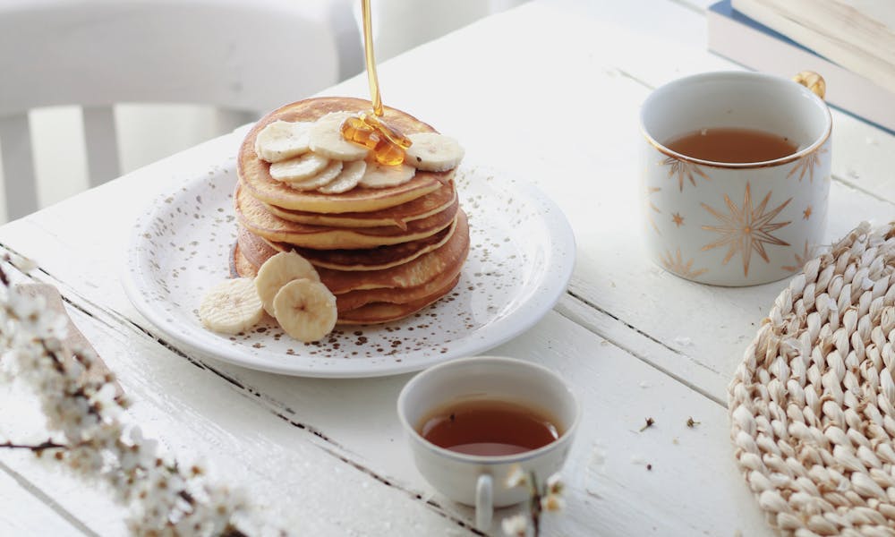 High angle of unrecognizable person adding caramel topping on tasty pancakes with banana slices served on table with cups of tea in morning .pexels