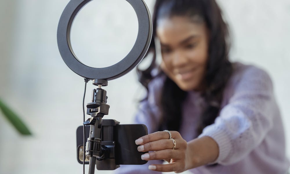 Cheerful young African American female blogger in stylish sweater smiling while setting up camera of smartphone attached to tripod with ring light before recording vlog .pexels