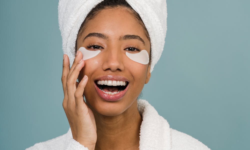 Cheerful young black female in white bathrobe with towel on head and eye patches laughing and looking at camera during skin care routine after shower .pexels