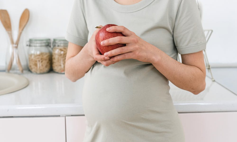 Unrecognizable pregnant female with ripe red apple for healthy diet in hands standing near counter in light kitchen at home .pexels