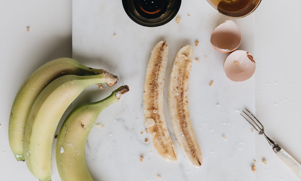 Top view composition of cut ripe bananas and bowl of raw egg on white marble board decorated with broken eggshell and fork .pexels
