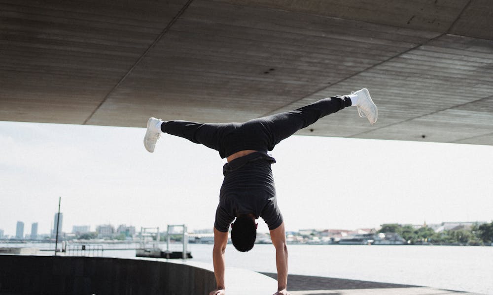 Back view full body strong sportsman performing handstand and split in air while working out on sunny city embankment .pexels