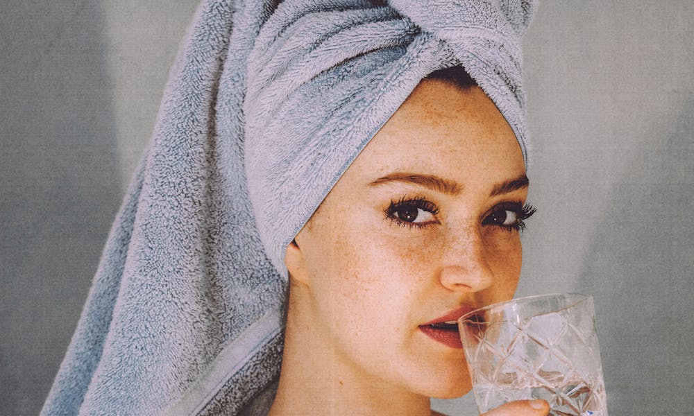 Young woman with hair wrapped with towel and bared shoulders drinking water and looking at camera .pexels