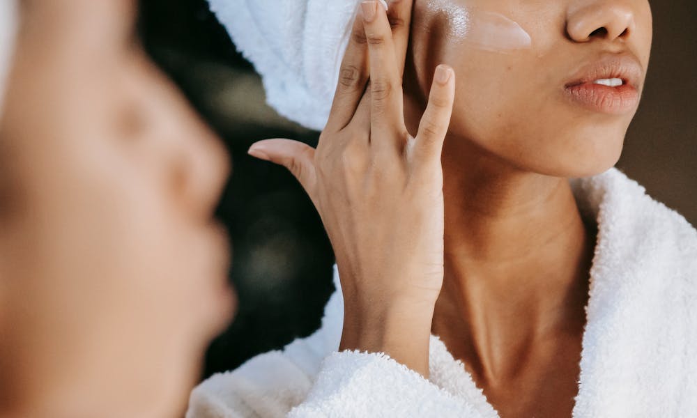 Crop unrecognizable young ethnic female in terry robe applying moisturizing cream on cheek while reflecting in mirror .pexels