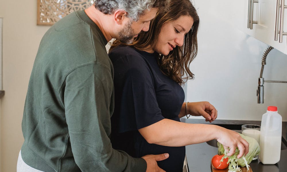 Side view of tranquil man in casual clothes embracing pregnant wife cooking salad in cozy kitchen in daytime .pexels