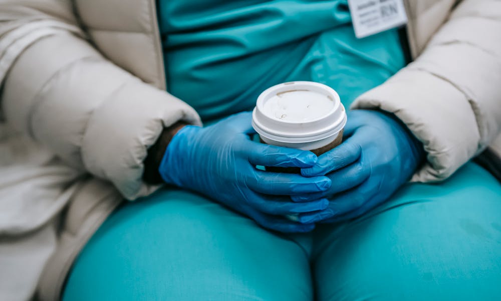 Crop anonymous plump nurse in uniform under warm wear and protective latex gloves sitting in public place and holding takeaway cup .pexels