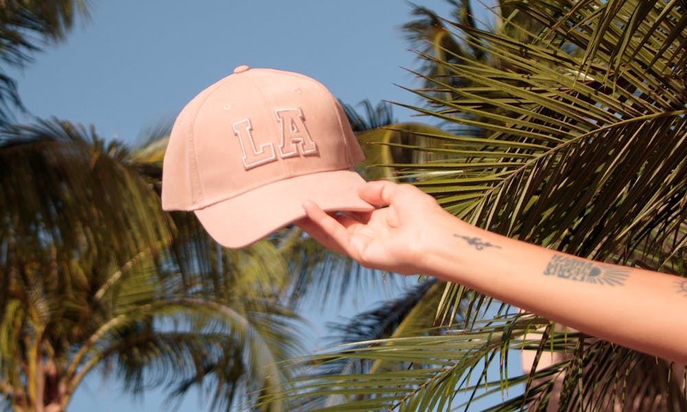 A pink LA cap is held up against a backdrop of palm trees under a clear blue sky in Los Angeles. .pexels