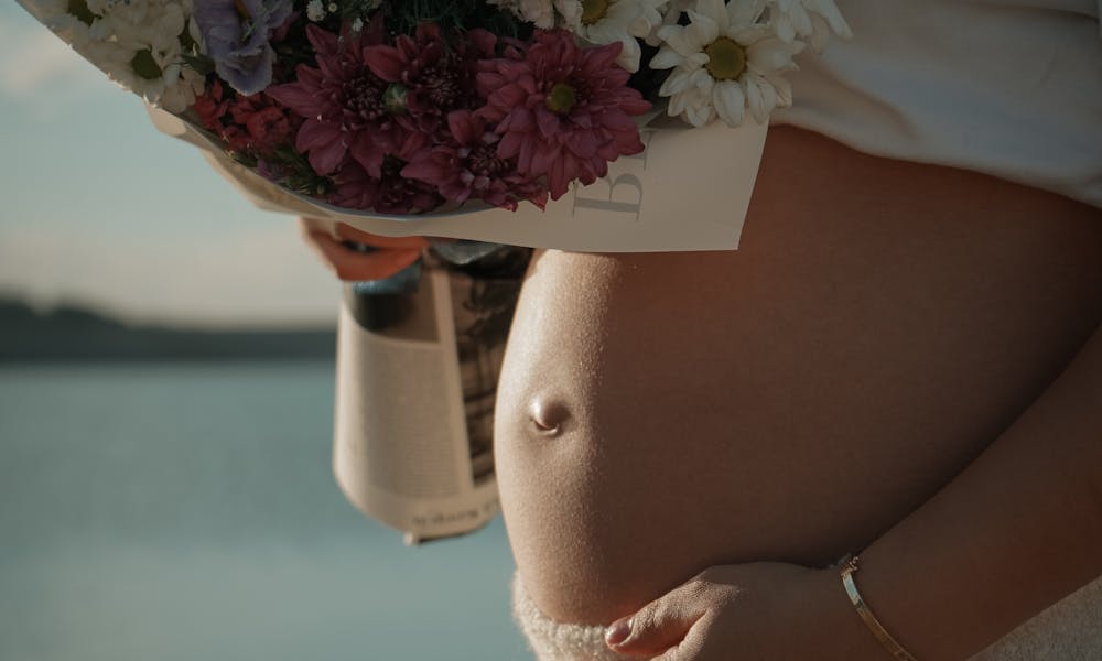 Pregnant woman cradling her belly and holding a bouquet of flowers by a serene lake. .pexels