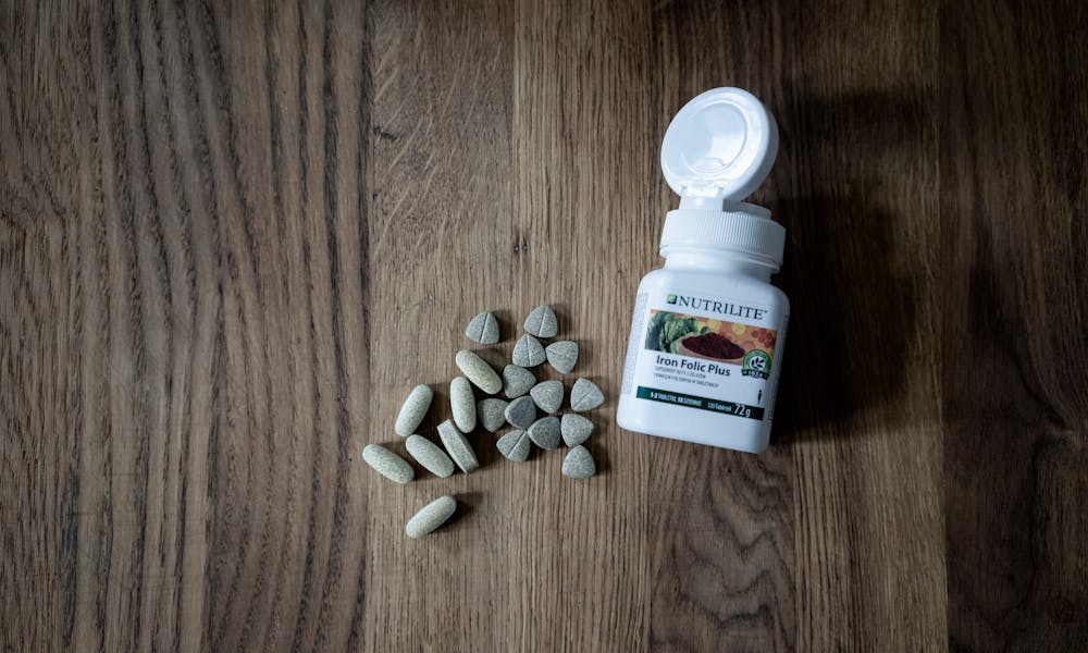 Open bottle of Iron Folic Plus tablets with scattered pills on a wooden surface. .pexels