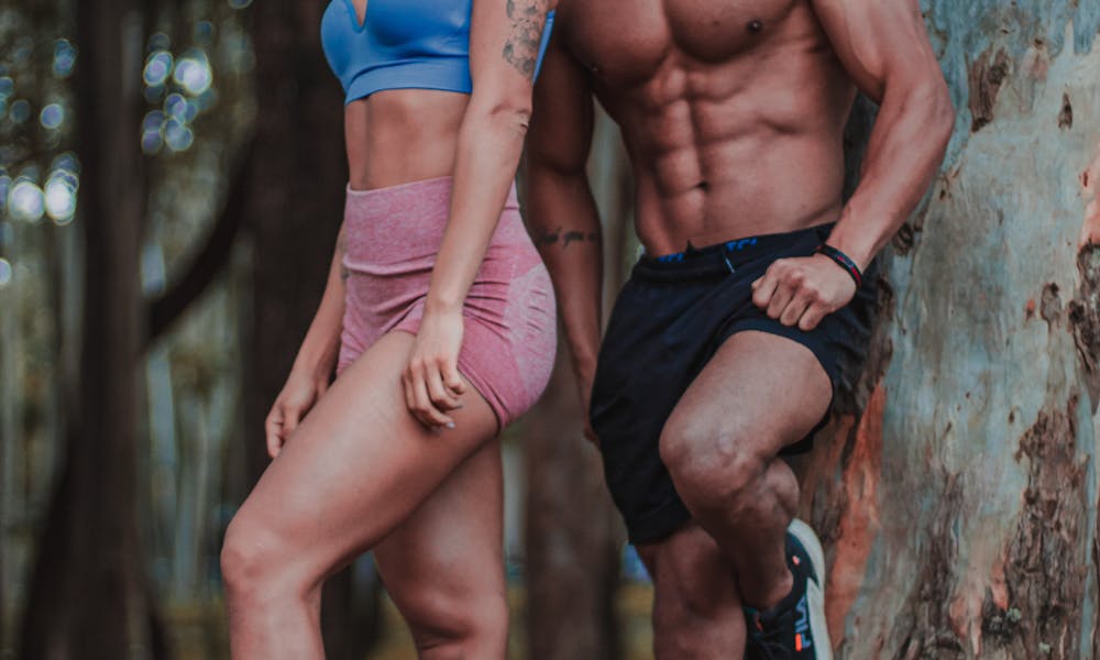 A fit couple posing confidently by a tree in a forest setting. .pexels