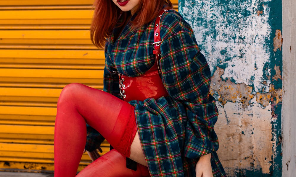 Chic red-haired woman posing in vibrant red and plaid attire against urban backdrop. .pexels