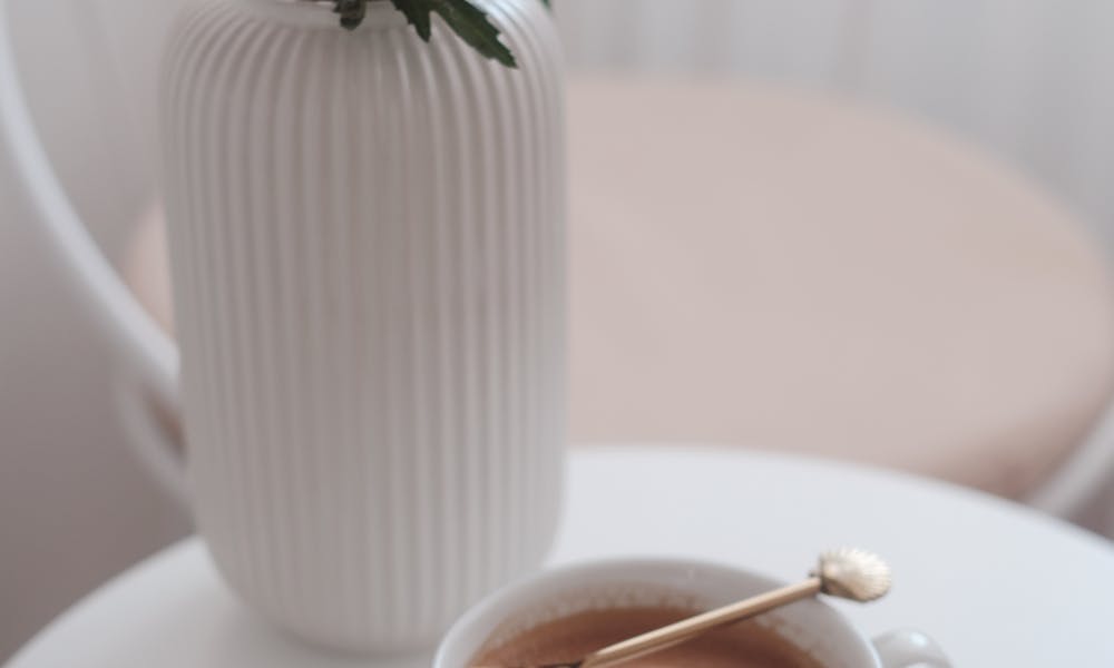 A cup of coffee with a gold spoon next to pink flowers in a vase on a white table. .pexels